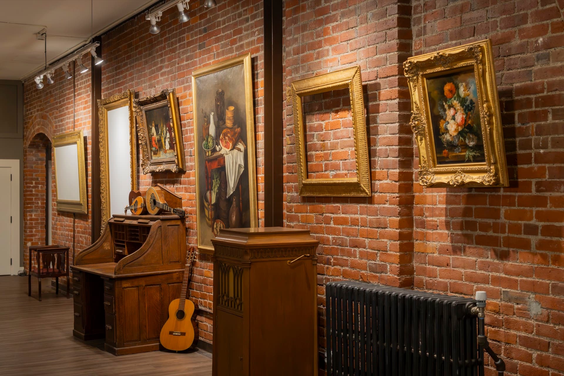 A second view of the Occidental gallery: framed contemporary and historical paintings on exposed brick, antique cabinet and acoustic guitar in the foreground.